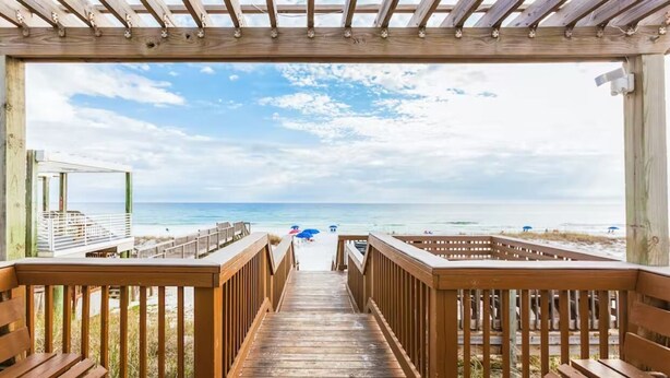 Un muelle de madera que conduce a una playa, con vista despejada del océano y bañistas a lo lejos.