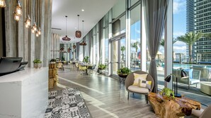 A modern hotel lobby with a long reception desk, multiple seating areas, and large windows offering views of the city and a pool.