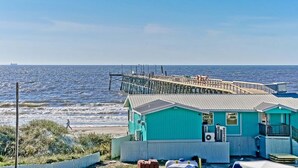 Ein Küstengebäude mit einem Steg, der ins Meer führt, ein Strand im Hintergrund und ein klarer Himmel.