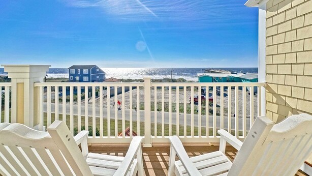Ein Balkon mit weißen Stühlen und Blick auf das Meer, sowie ein Gebäude mit sandfarbener Fassade.