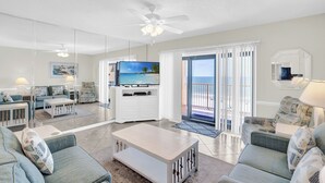 Ein geräumiges Wohnzimmer mit großem Flachbildfernseher, bequemen Sitzgelegenheiten, einem Deckenventilator und Blick auf den Strand durch eine Glastür.