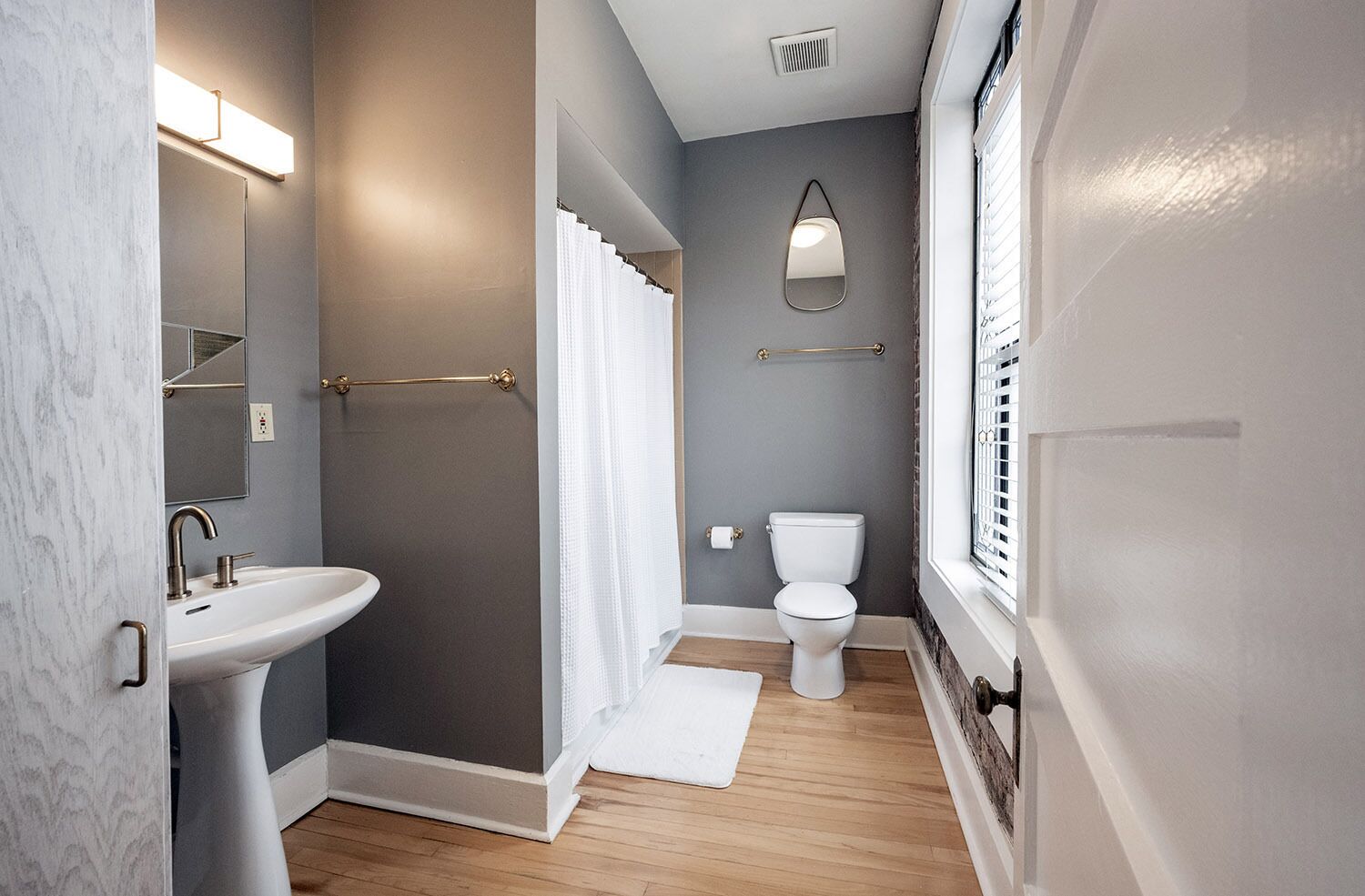 A narrow bathroom with a pedestal sink, a shower with a curtain, a toilet, and a mirror.