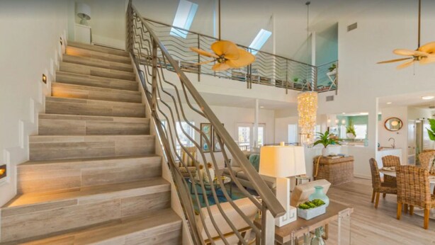 A spacious living area with a wooden staircase, a dining area with wicker furniture, and a ceiling fan. - Sandcastle Beach House, Ocean Views, 5Bed 4.5Bath, Huge Outdoor Private Pool (Port Aransas,)