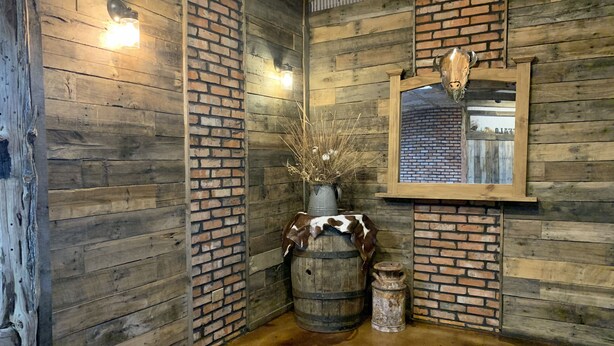 Ein rustikales Interieur mit Holzwänden, einer Wand aus Ziegelsteinen, einem Holzfass, einem Kuhfell und einer Vase mit Trockenpflanzen.
