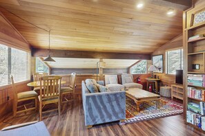 Cozy cabin with a high ceiling and exposed beams, featuring a seating area with a sofa and chairs, dining table, and bookshelf.