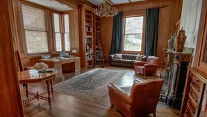 A spacious living room with a wooden floor, a large bookshelf, a sofa, a desk with a laptop, and a fireplace. - Mt Vernon Villa (Waipukurau)