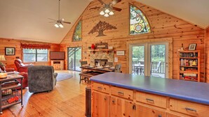 A spacious living room with wooden walls, a stone fireplace, a large dining table, and a ceiling fan. - 'River's Edge' - Ultimate Riverfront Getaway (Hayesville)