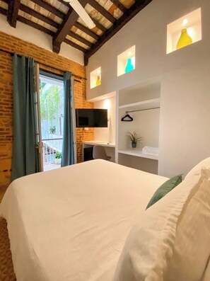 Una habitación de hotel con una cama grande, un televisor montado en la pared, una pequeña estantería con una planta y un balcón con cortinas.
