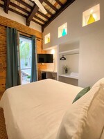 Una habitación de hotel con una cama grande, un televisor montado en la pared, una pequeña estantería con una planta y un balcón con cortinas.