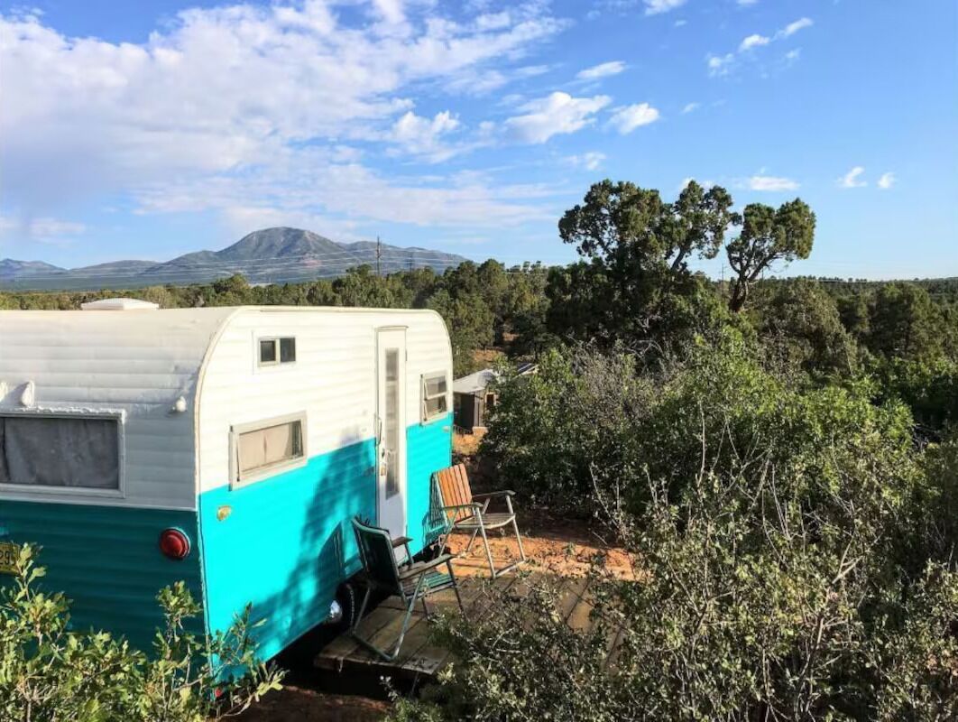 A white and turquoise RV parked in a natural setting with a chair and a table nearby.