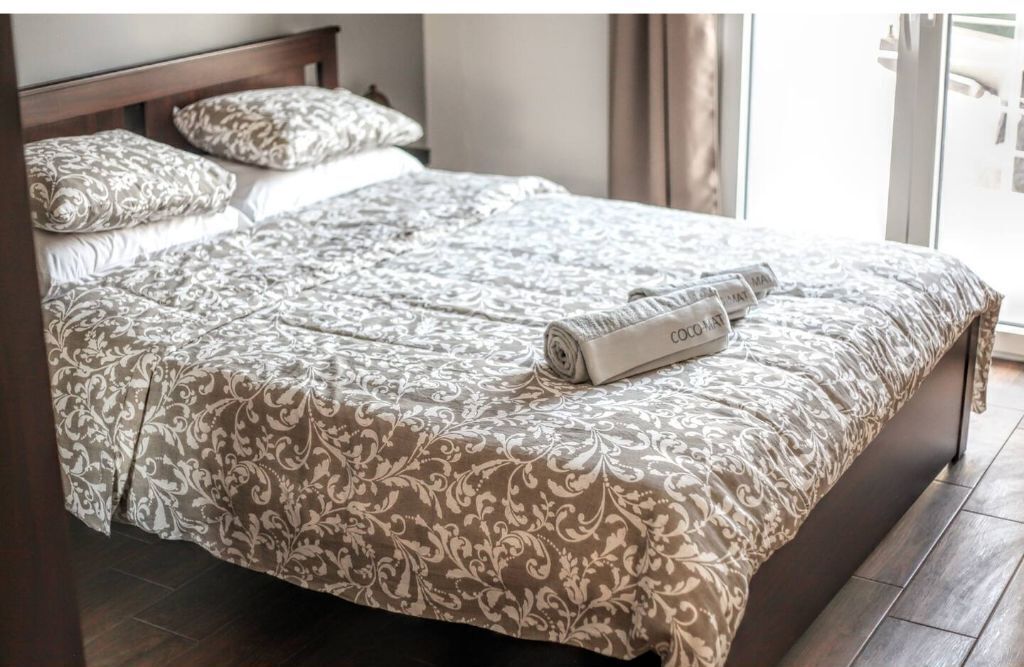 Ein ordentlich gemachtes Bett mit einem gemusterten Bettbezug und Kissen, ein Holzkopfstück und ein Fenster mit Vorhängen.