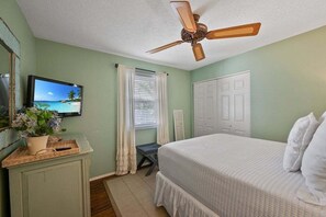 Une chambre à coucher avec un lit, un ventilateur de plafond, une télévision, une fenêtre avec des rideaux et une porte.