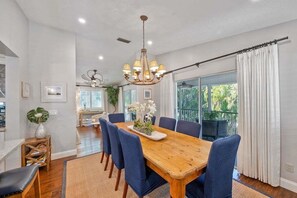 A dining area with a wooden table, blue chairs, a chandelier, and a view through large windows.