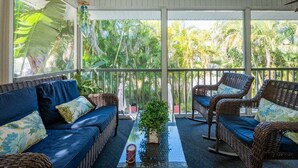 Un espacio de estar al aire libre con techo, con mobiliario de mimbre y una mesa de vidrio, con vistas a un jardín tropical.
