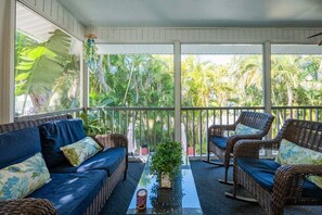 A covered outdoor seating area with wicker furniture and a glass table, overlooking a tropical garden.