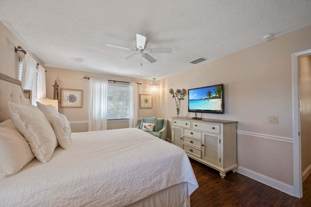 A bedroom with a bed, a ceiling fan, a television, a dresser, and a window with blinds.