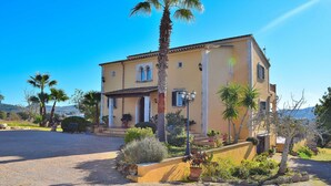 A two-story building with a yellow facade, a driveway, and surrounding greenery including palm trees and shrubs. - Can Claret Gran 176 wonderful villa with private pool, large terrace, air conditioning and WiFi (Cas Concos)