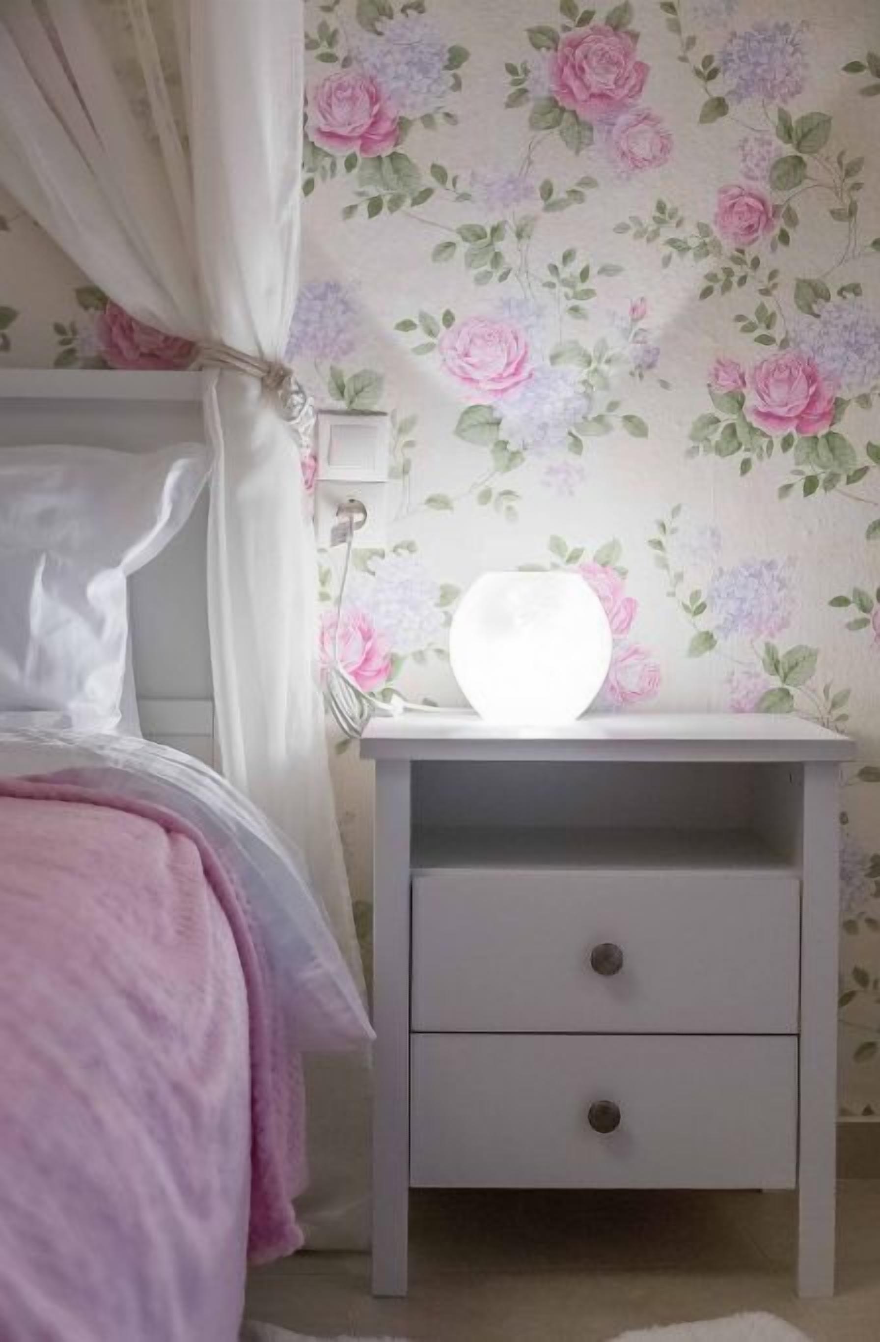 A bedroom with a bed, a bedside table with a lamp, and floral wallpaper.