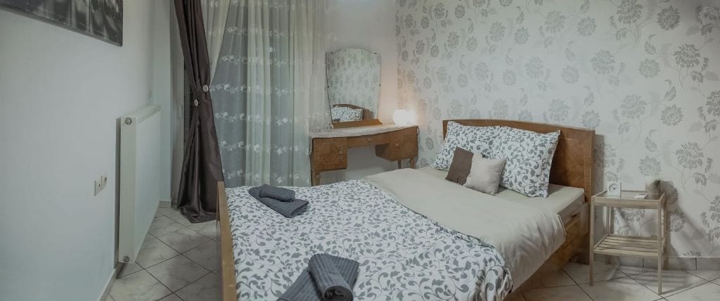 A bedroom with a bed, a bedside table, a chair, a mirror, and a window with curtains.