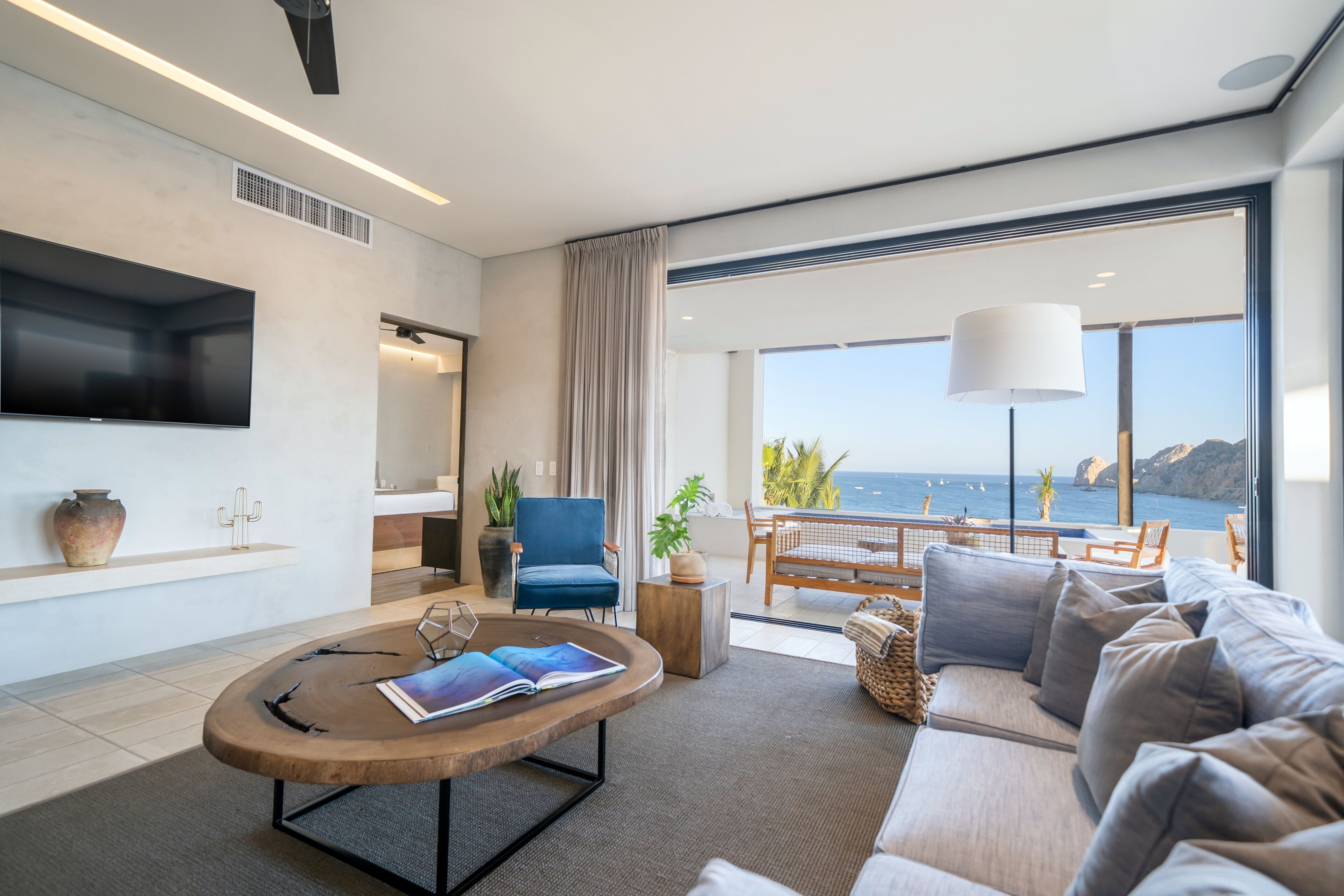 A modern living room with a large sofa, a coffee table, and a TV mounted on the wall. There is a view of the ocean through large windows.