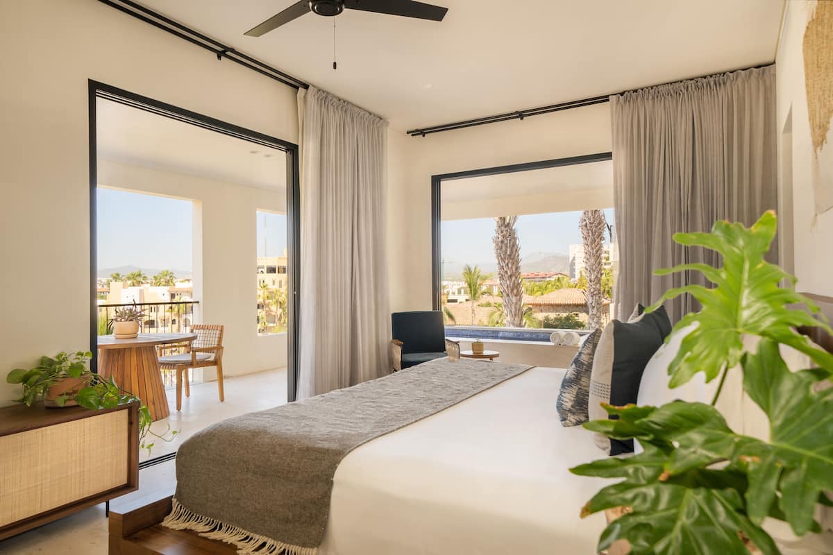 A modern hotel room with a large bed, a balcony with a table and chairs, and a view of the cityscape.
