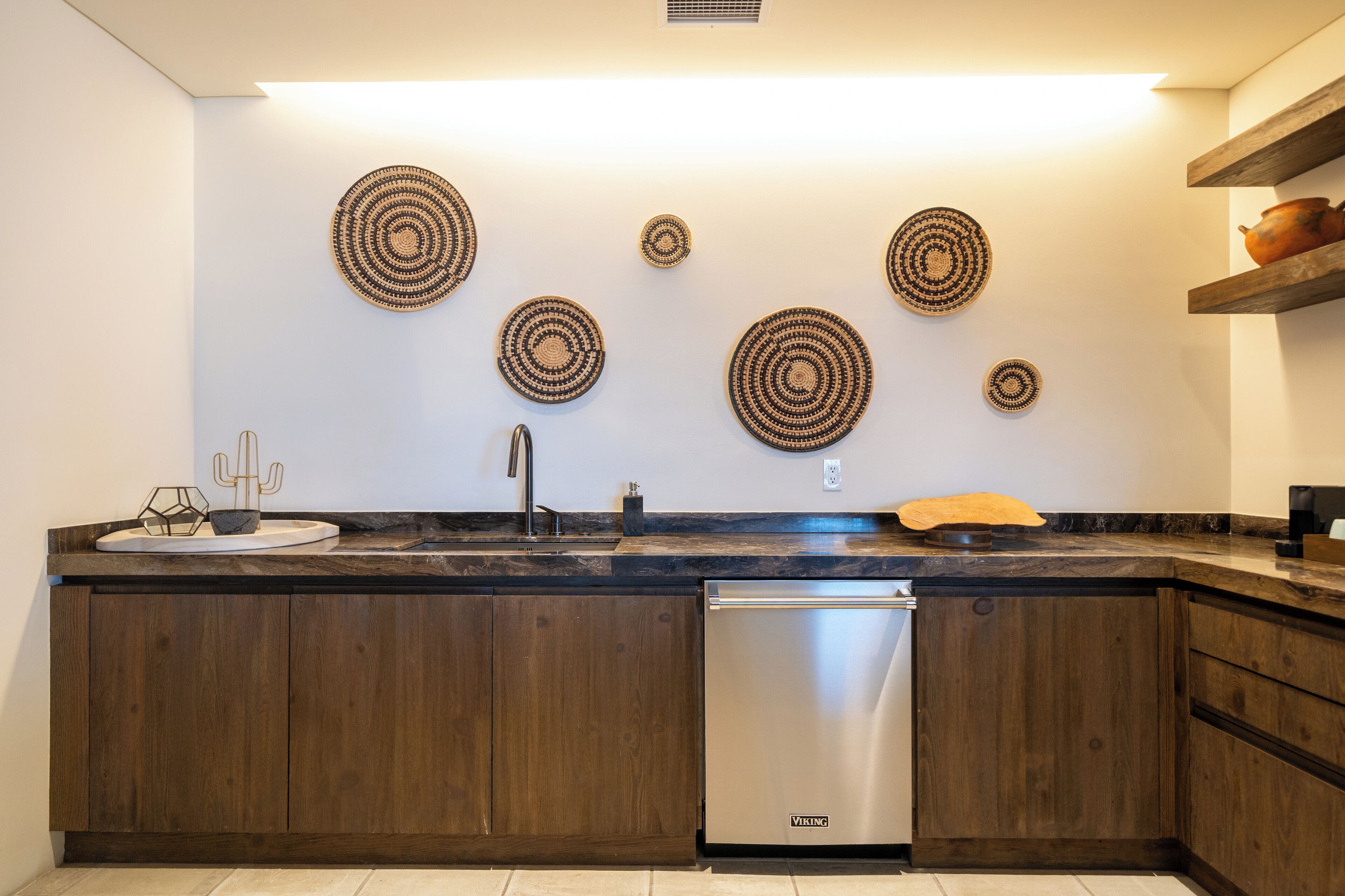 A modern kitchen with wooden cabinets, a stainless steel dishwasher, and decorative woven wall art.