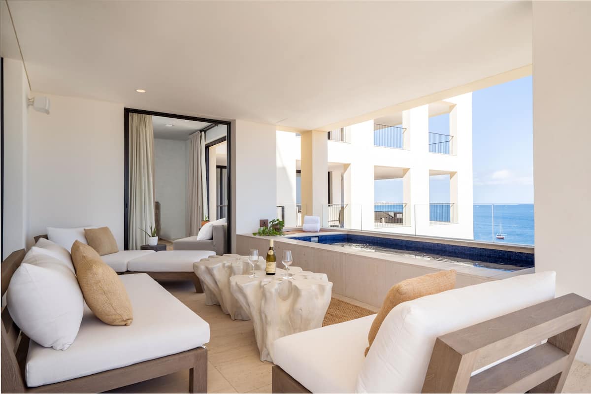 A modern hotel room with a balcony, white furniture, a pool, and a view of the sea.
