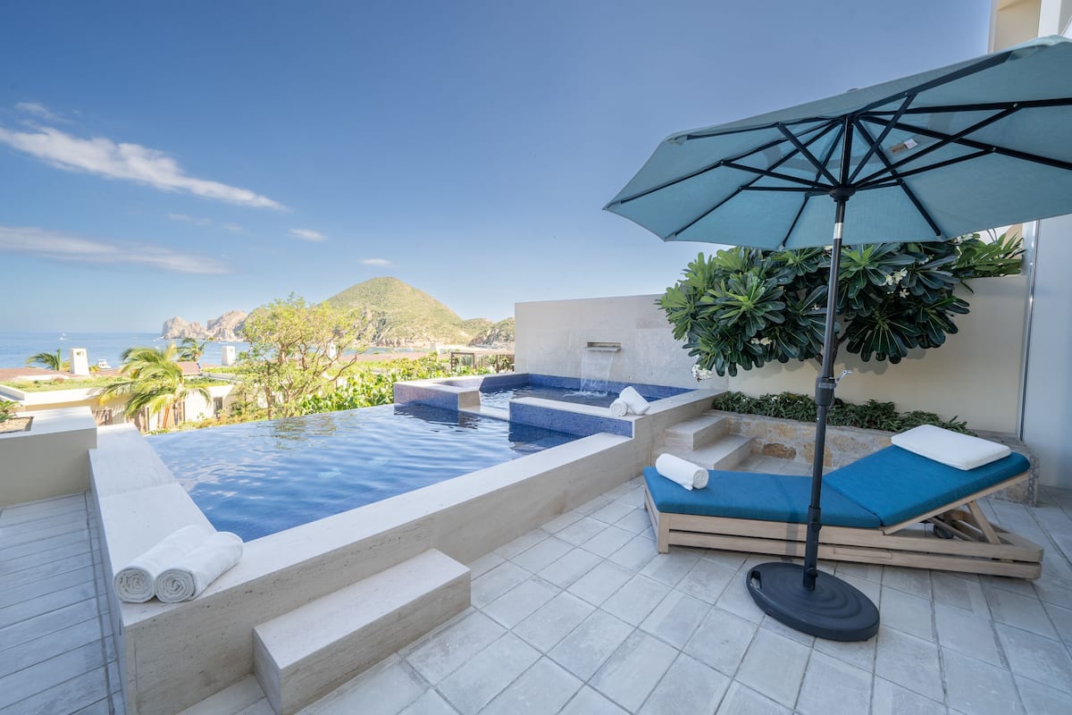 A rooftop infinity pool with a view of the sea, a lounge area with a blue cushion, and a large umbrella providing shade.