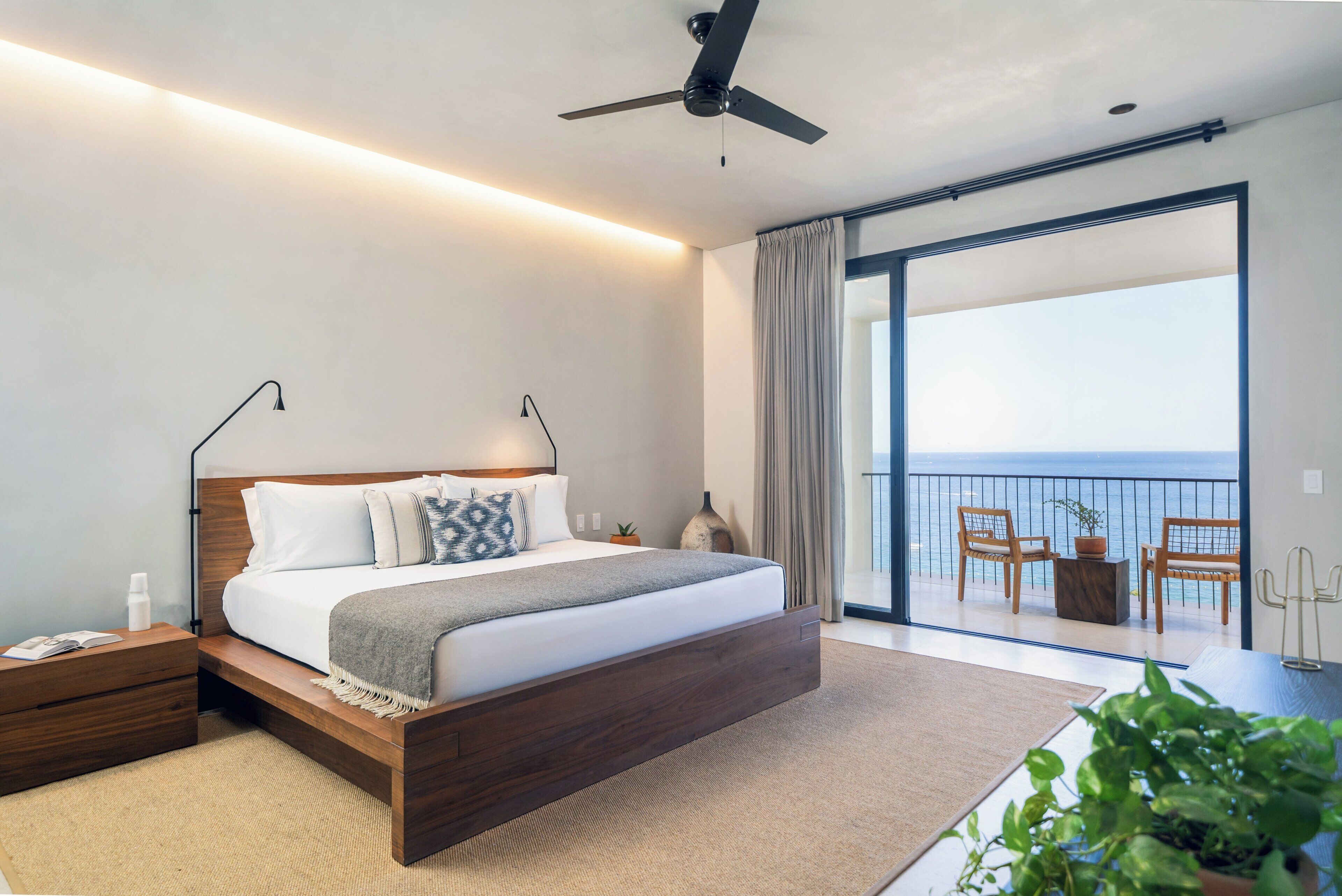 Un dormitorio moderno con cama, mesitas de noche, ventilador de techo y balcón con sillas y vistas.