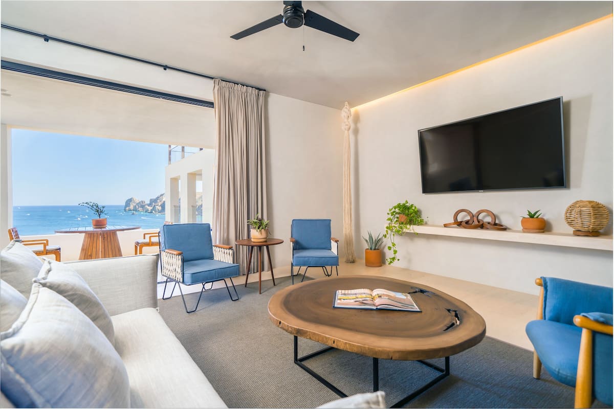 A modern living room with a large window overlooking the ocean, a flat-screen TV mounted on the wall, and a ceiling fan.