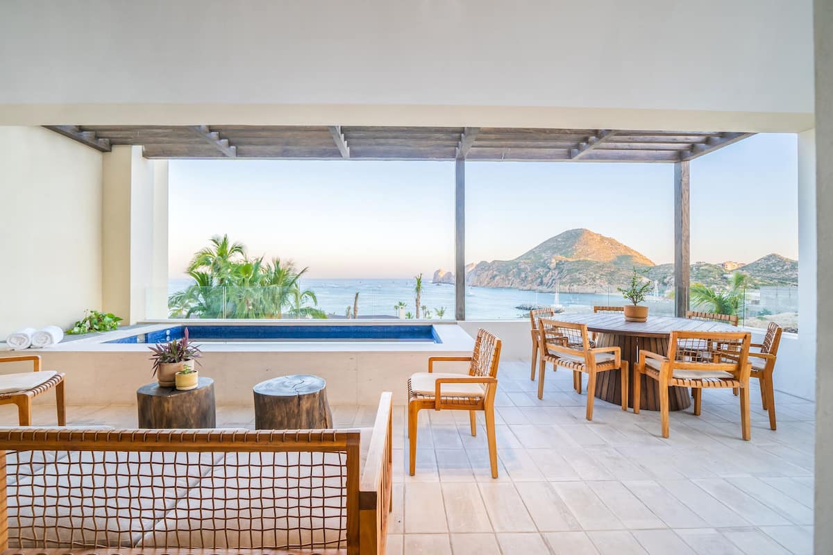 A terrace with a pool, wooden furniture, and a view of the sea and a mountain.