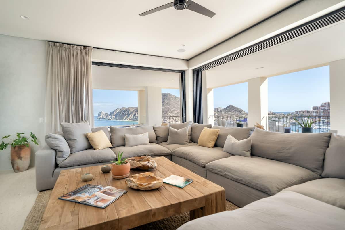 A modern living room with a large sectional sofa, a wooden coffee table, and a view of the sea and mountains.