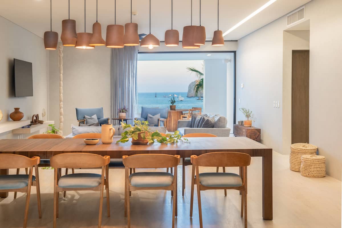 A modern dining area with a wooden table, four chairs, hanging pendant lights, and a view of the sea through large windows.