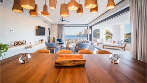 Un comedor moderno con una mesa de madera, sillas color naranja, lámparas colgantes y vistas al océano.