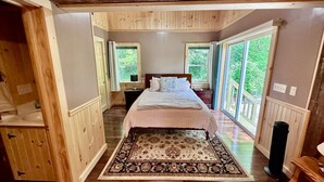 A bedroom with a bed, a wooden floor, a patterned rug, and large windows.