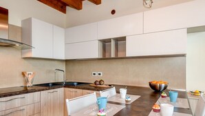 Eine moderne Küche mit weißen Schränken, einer Edelstahlspüle und einer Holzausziehtafel. Zwei Stühle und ein Tisch mit Platzdeckchen sind vorhanden.