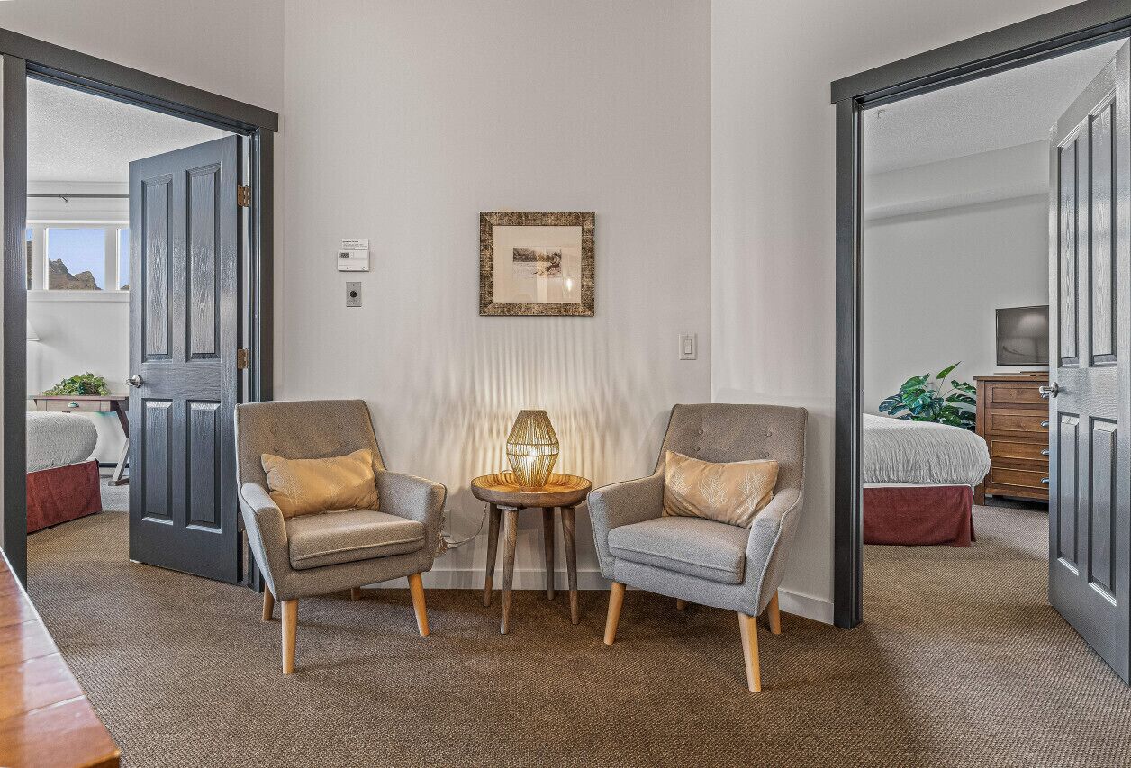 A room with two armchairs, a small table with a lamp, a framed picture on the wall, and a bed in the background.