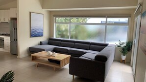A modern living room with a grey sectional sofa, a wooden coffee table, and a large window offering a view of greenery. - Contemporary On Cuttriss - Inlet Side (Inverloch)