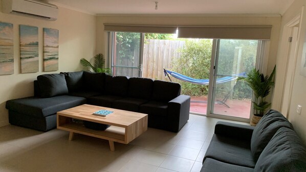A living room with a sectional sofa, a wooden coffee table, and a large sliding glass door leading to a backyard with a hammock. - Contemporary On Cuttriss - Inlet Side (Inverloch)