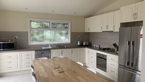A modern kitchen with white cabinets, a wooden dining table, stainless steel appliances, and a window with a view of trees. - Contemporary On Cuttriss - Inlet Side (Inverloch)