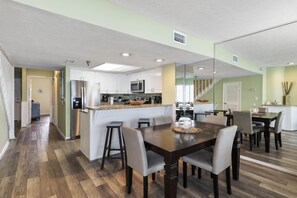 A modern kitchen with white cabinets, a stainless steel refrigerator, and a microwave. There is a dining area with a wooden table and chairs. - GULF FRONT PARADISE! 201 Destin Seafarer (Destin)