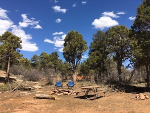 A campsite with two chairs, a picnic table, and a fire pit surrounded by trees and dry ground.