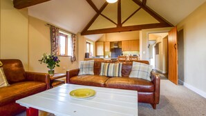 A cozy living room with a brown leather sofa, a white coffee table, and a vase of flowers. A kitchen is visible in the background. - Cider House at Oosland Barns - sleeps 4 guests  in 2 bedrooms (Bidford-on-Avon)