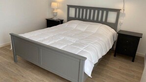 A neatly made bed with a grey headboard and footboard, a bedside table with a lamp, and a small drawer. - Nice apartment 4 people in Hennebont wifi -  near Lorient 3 stars (HENNEBONT)