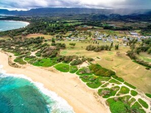 An aerial view of a coastal area with a beach, green vegetation, and a small settlement with houses. - Idyllic 90 Day North Shore Getaway! (Kahuku)