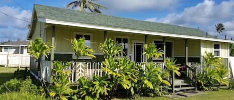 A single-story house with a green roof, a porch with a white railing, and a small front yard with tropical plants.