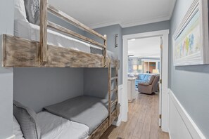 Habitación con literas de madera, un escritorio pequeño y una silla. Hay una puerta que da a otra habitación con un sofá y una mesa.