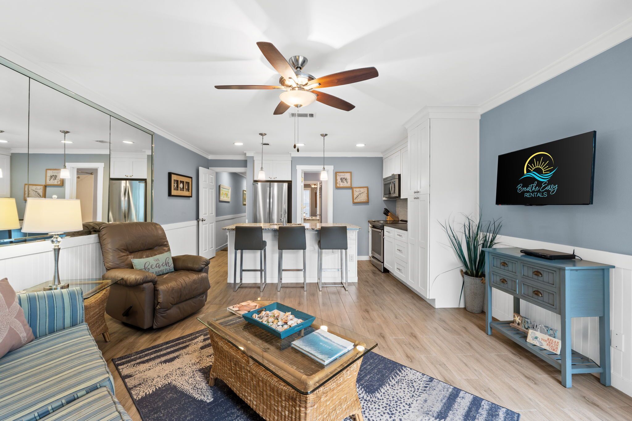 A modern living room with a brown leather chair, a glass coffee table, and a blue sofa. There is a kitchen area with white cabinets and a bar stool.
