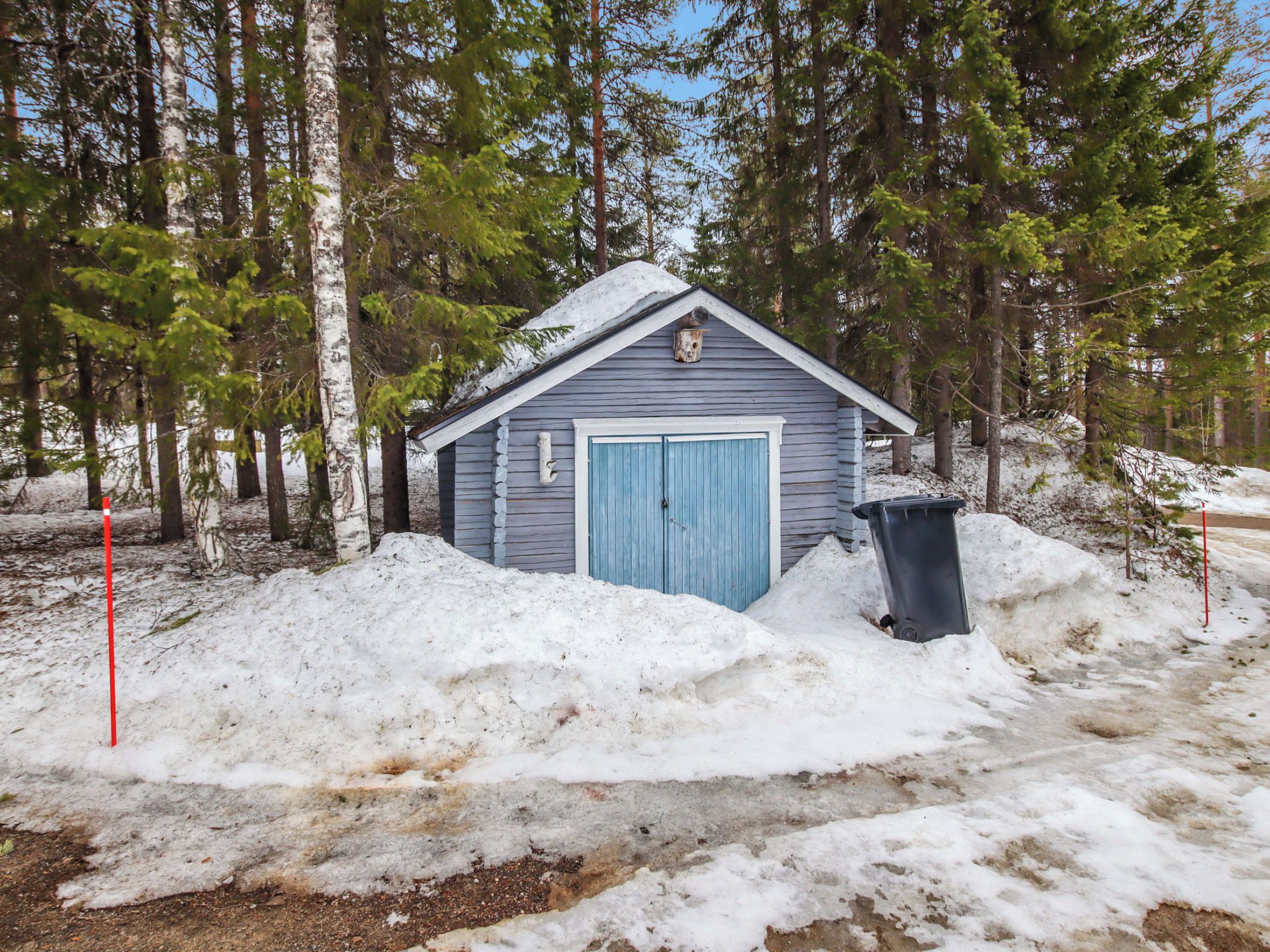 A small, single-story cabin with a blue door, surrounded by snow and trees.