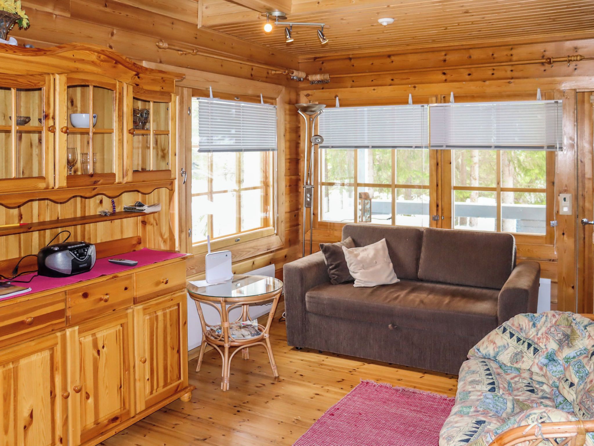 Interior de una cabaña de madera con kitchenette, sofá, mesa de comedor y ventanas amplias.